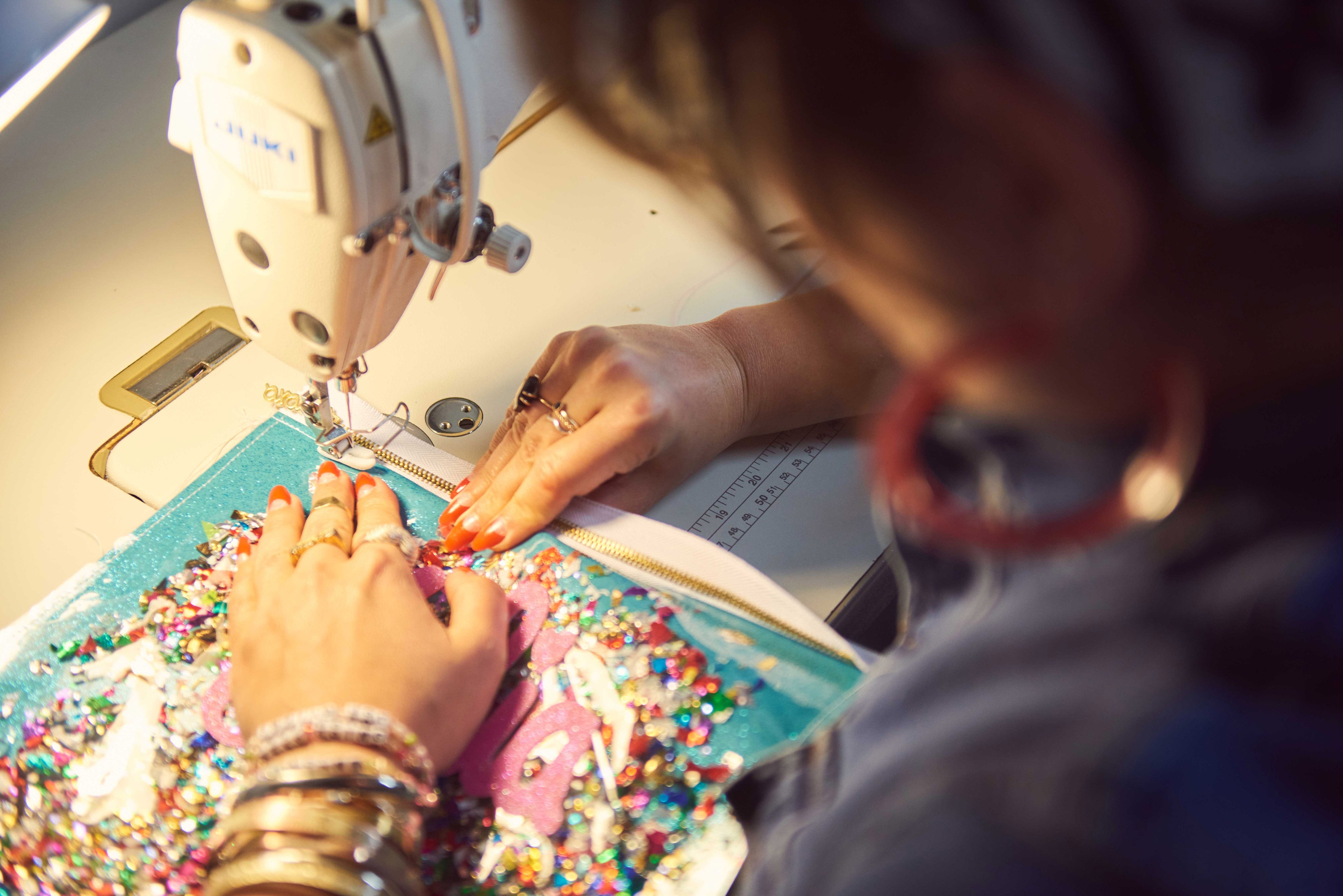 julie mollo sews on an industrial juki machine making a custom clutch that has confetti inside it. clutch is blue glitter vinyl with rainbow confetti on top and the name julia in pink glitter canvas