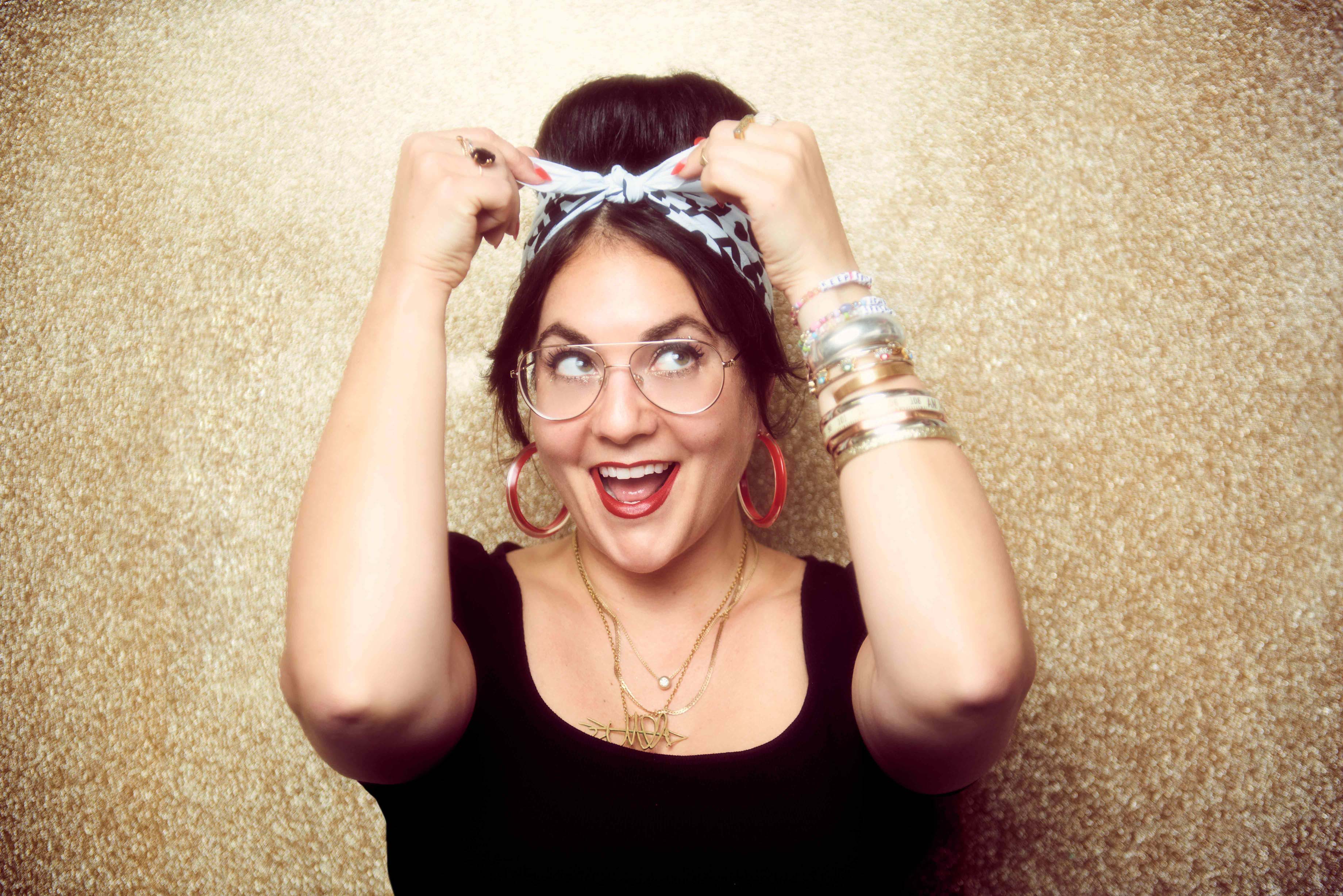 retro looking woman on a gold glitter background in a black top and gold chains. black and white doodle printed bandana is tied around her head and she has red hoop earrings and a stack of bracelets on