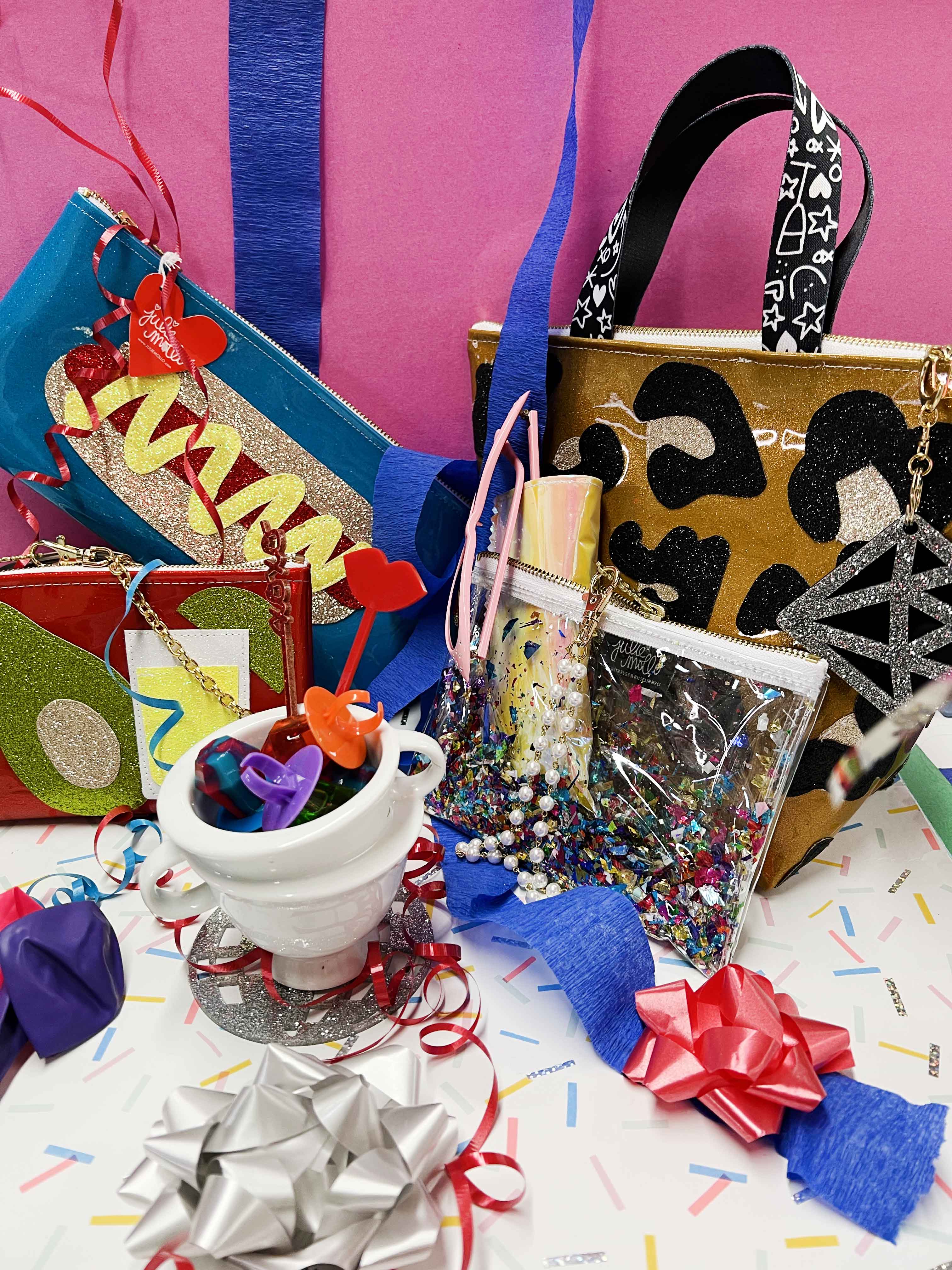 Vibrant display of Julie Mollo accessories against a pink background. Features a glitter hot dog-themed clutch, a red martini clutch, a leopard-print 'Small Business Bag' tote, and clear pouches filled with confetti. Surrounding decorations include colorful streamers, red and silver bows, drink stirrers in a cup, pearls, and festive ribbons, creating a lively celebration theme.