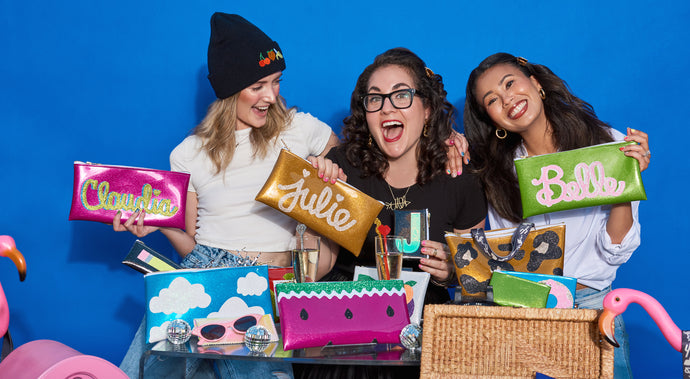 three girls hold custom clutches with their names on them on a blue background surrounded by glitter vinyl clutches with shapes. custom clutches read Claudia with hot pink glitter vinyl and green and blue writing, gold vinyl clutch with Julie written in gold, Lime green vinyl clutch with pink and white glitter canvas writing