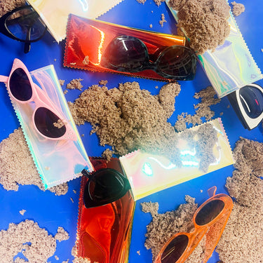 Playful product display for iridescent sunglasses sleeves with sand piles on a blue background with different colored sunglasses on top of and coming out of the various sunglasses sleeves. 