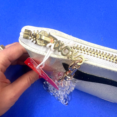 A close-up of a white clutch with a gold XOXO zipper pull. Connected to the zipper is a clear acrylic chain wrist strap.
